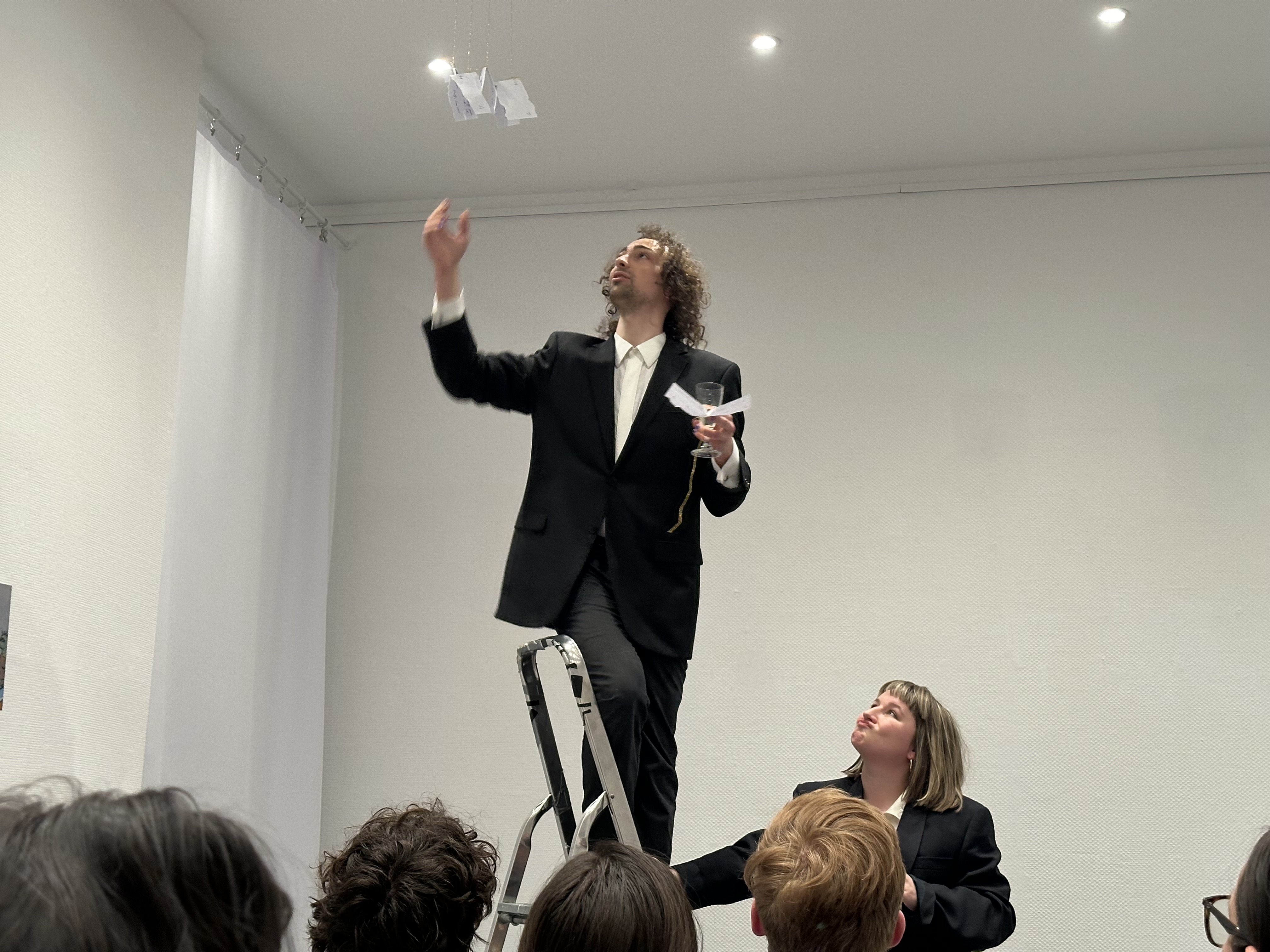 Suited Dimitri and Ioana pull slips of paper from the ceiling of the whitewalled gallery